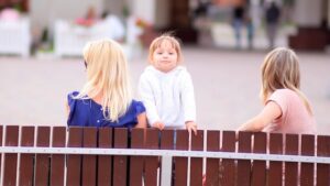 Kind steht auf Bank zwischen zwei Frauen und schaut in die Kamera
