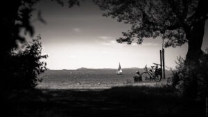 Ein Mensch sitzt neben Fahrrädern vor See mit Segelschiff