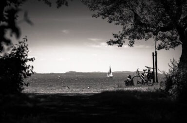Ein Mensch sitzt neben Fahrrädern vor See mit Segelschiff