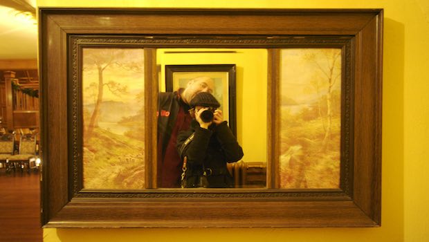 Paar steht vor Spiegel und gelber Wand, sie hält eine Kamera