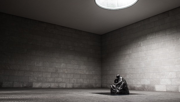Sitzende Skulptur im Raum vor Steinmauer, darüber eine Lampe