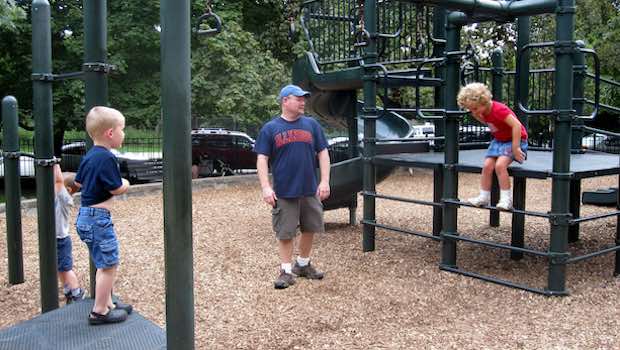 Vater steht auf dem Spielplatz beim Klettergerüst mit zwei Kindern